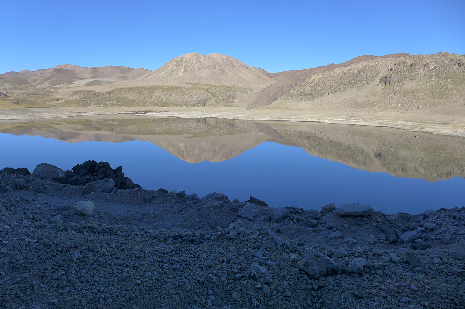 Stony, brown-gray dome reflected perfectly in cobalt blue lake, against clear blue sky.