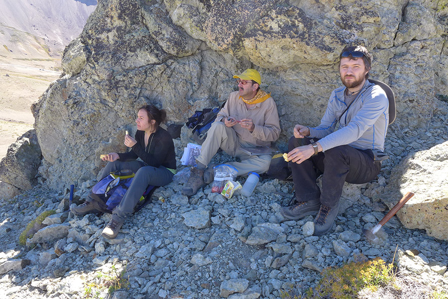 3 people in shade of large rock look out on volcanic landscape, 2 rock hammers are visible.