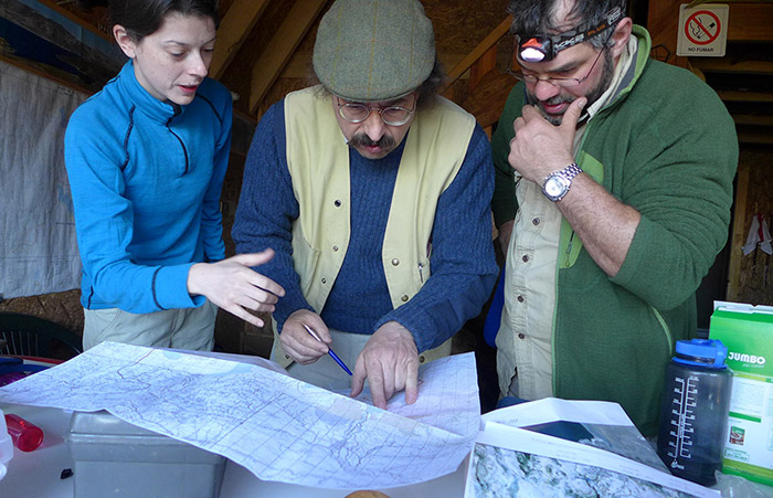 Woman and 2 men pore over map inside cabin, with puzzled expressions.