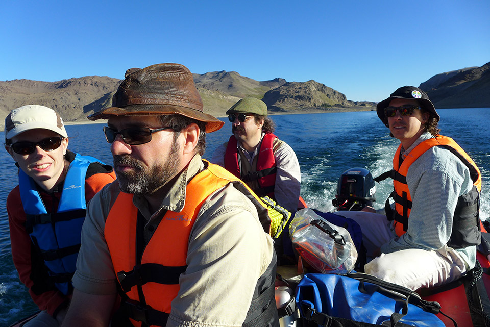 4 people, all with hats and sunglasses, look forward as a boat crosses a lake on a bright, sunny day.