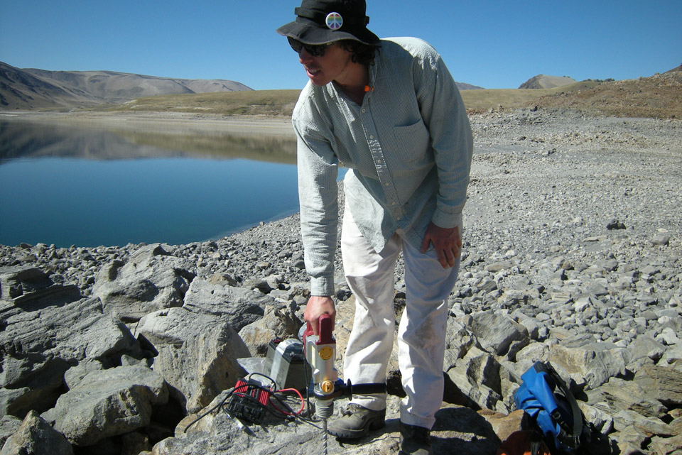 Man holds drill against gray rock; banded gray rocks in foreground, lake in background.
