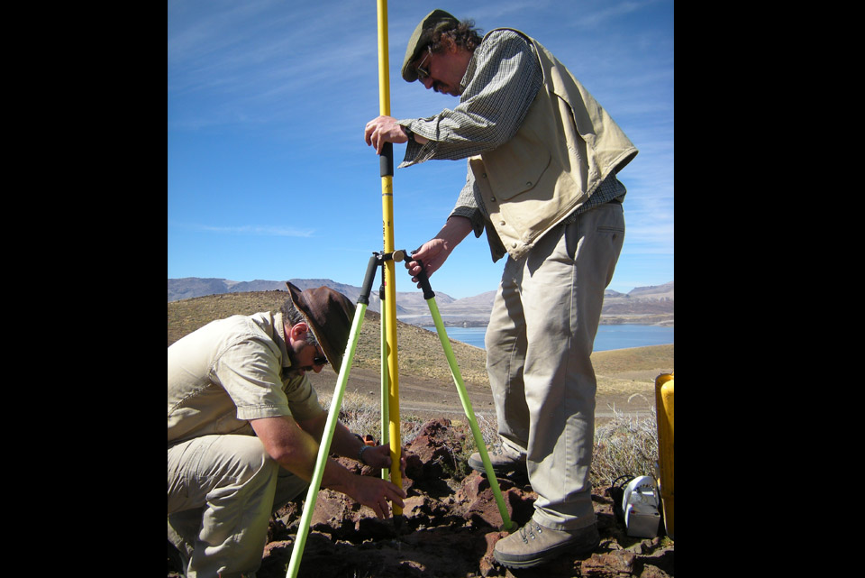 2 men hold a tripod for the GPS unit. 1 of them places a yellow vertical pole on top of a large red rock. Blue sky with wispy clouds.