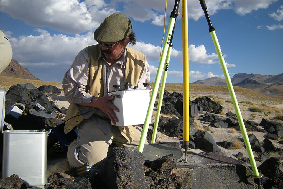 Man holds rectangular white case as he pivots toward a rock holding GPS tripod. Black boulders litter the landscape.