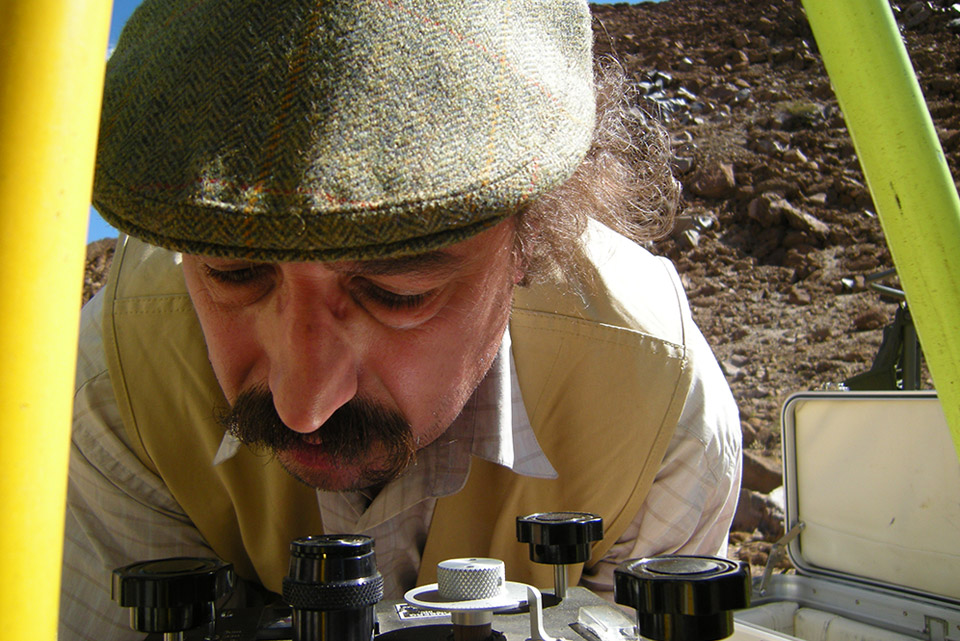 Man wearing cap peers intently through a lens on instrument covered with dials. Photo framed by yellow legs of GPS tripod.