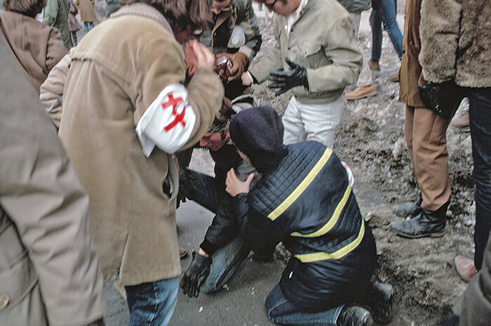 Medic with medical red and white armband assisting an injured male protestor on the ground outdoors. Photo by John Wolf