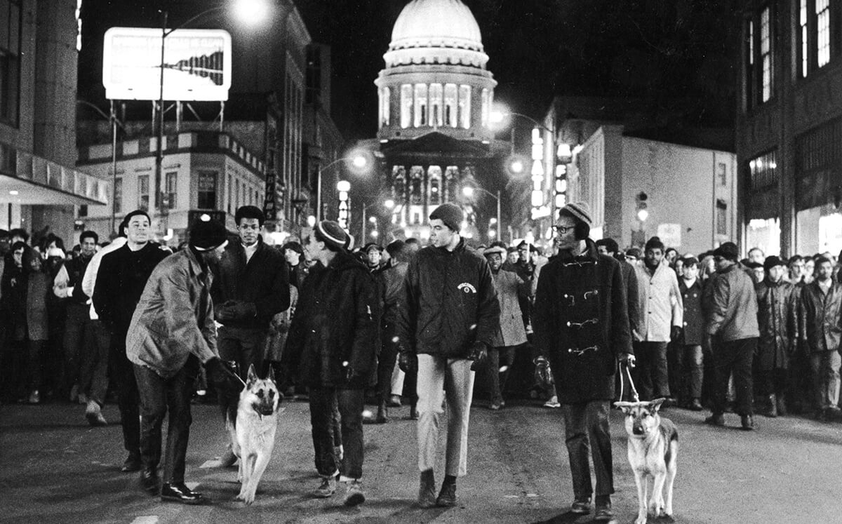 Five African American campus leaders lead the nighttime march down State Street with German Shepherd dogs and the Wisconsin State Capitol lit up in the background. Photo: Wisconsin State Journal