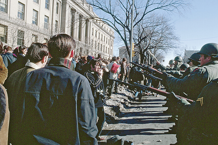 National Guardsmen lined up outdoors and point their bayonets toward protesting students, one of them standing directly in front of them taking a picture. Photo by John Wolf