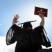 Person in cap and gown holding up diploma cover in the sunshine