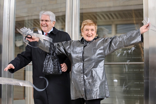 Photo of Simona Chazen, flinging her arms wide in celebration, as she and husband Jerome cut a giant silver ribbon.