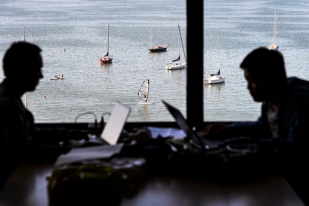 The juxtaposition of campus life during summer session is seen as two silhouetted students study indoors next to a lakefront window in College Library at Helen C. White Hall. Outside in the background, paddleboarders, docked boats and a lone windsurfer dot Lake Mendota.
