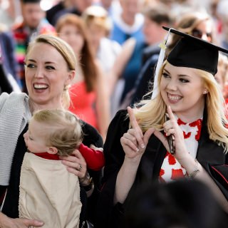 Soon-to-be graduate Alexa Price flashes a W hand symbol as her mother, Bethany Lopresti, carries little brother, Xavier, while walking toward the start of UW-Madison's spring commencement ceremony at Camp Randall Stadium at the University of Wisconsin-Madison on May 13, 2017. The outdoor graduation is expected to be attended by more than 6,000 bachelor's and master's degree candidates, and their guests. (Photo by Jeff Miller/UW-Madison)