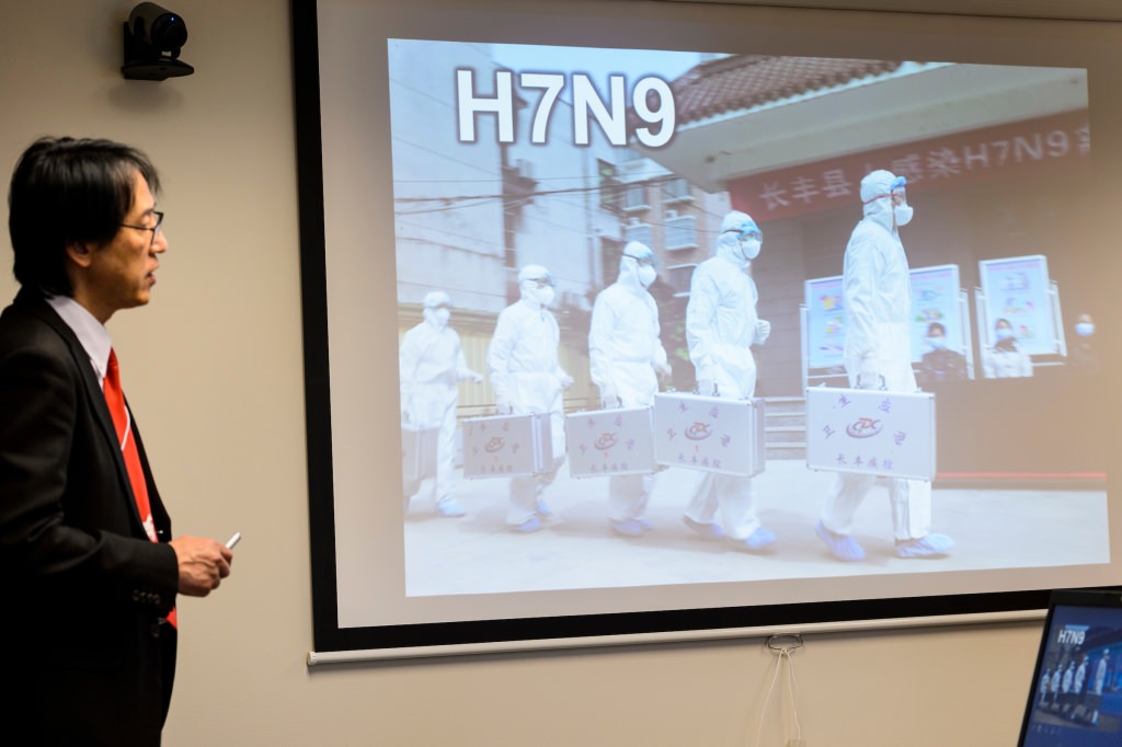 Photo: Kawaoka in front of screen projecting slide of people in hazmat suits
