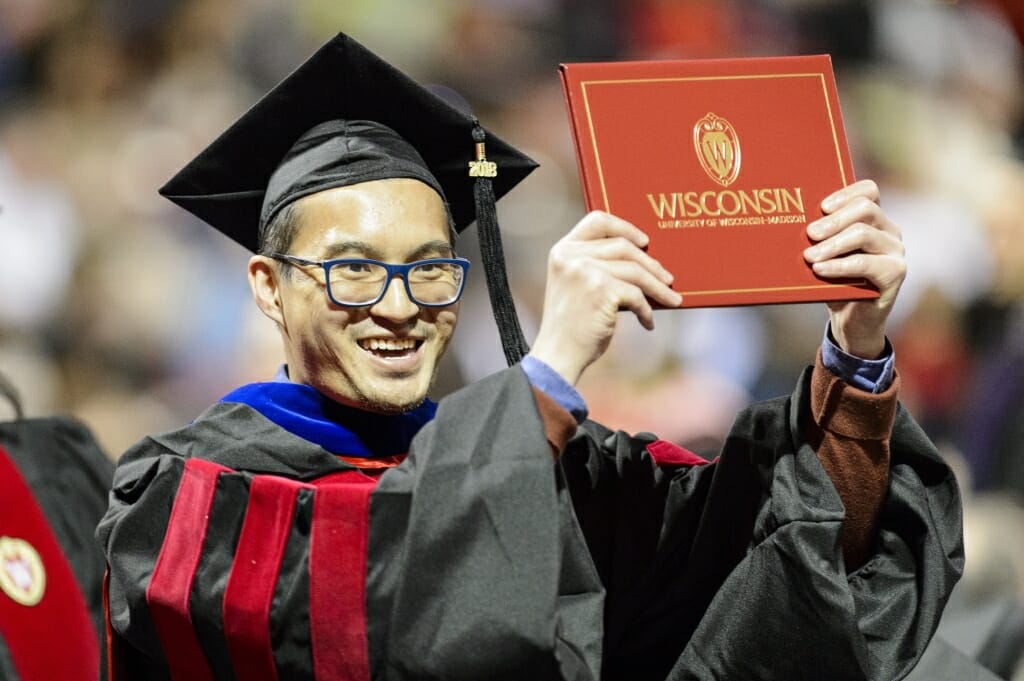 Photo of Khoa Tran, a PhD graduate in molecular and cellular pharmacology, showing off his diploma cover to his family in the stands.