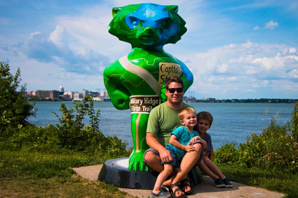 A man and his two young sons sit on a pedestal of a Bucky statue overlooking Lake Monona.