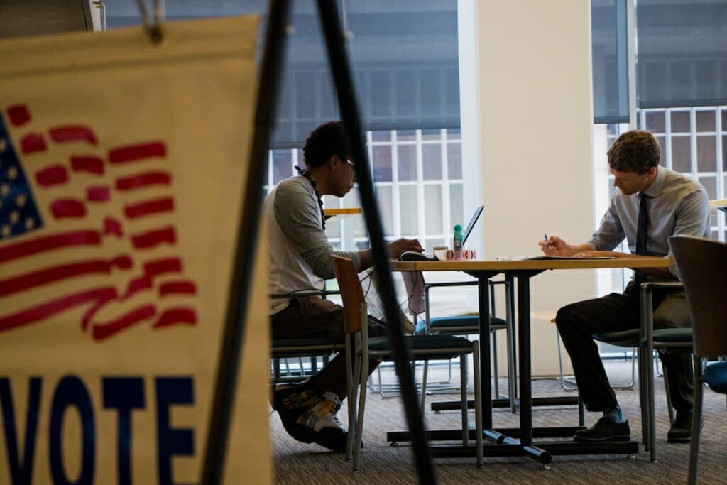 Photo: Poll worker and voter sitting at table with Vote sign in foreground