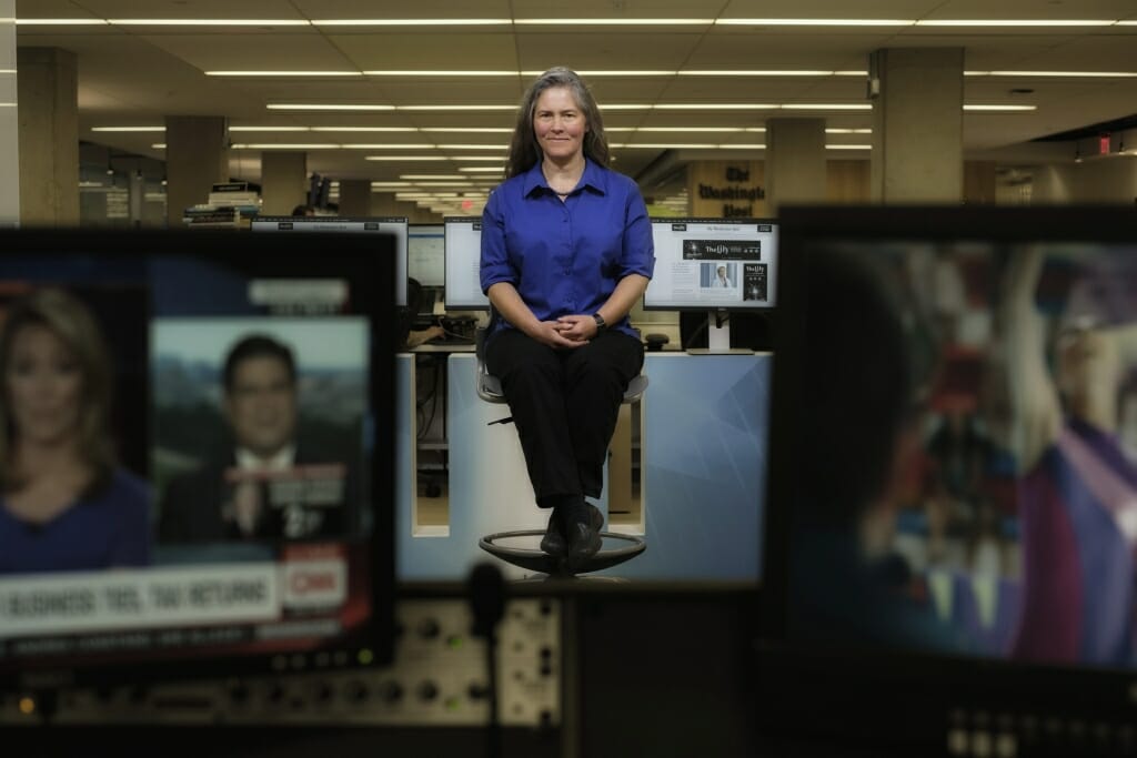 Photo: Laura Helmuth sitting on a stool between TV monitors showing news programs