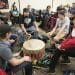 A number of students sit around a Native drum and bang it.