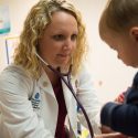 A physician, Hope Villard, has long, curly blond hair and is wearing a white doctor's coat. She has a stethoscope in her ears to listen to the heartbeat of her patient, a toddler, sitting on an exam table in a clinic setting.
