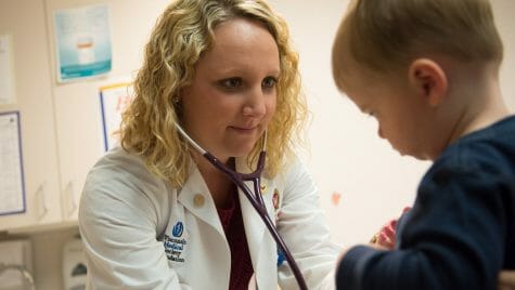 A physician, Hope Villard, has long, curly blond hair and is wearing a white doctor's coat. She has a stethoscope in her ears to listen to the heartbeat of her patient, a toddler, sitting on an exam table in a clinic setting.