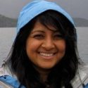 Photo: Apoorva Mandavilli standing in a hooded raincoat in front of a lake with mountain in background