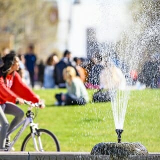Photo: A fountain is in the foreground, and students sit on the grass in the background. One student bicycles.