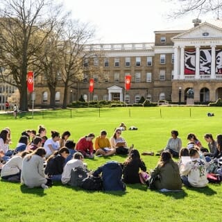 Photo: About 20 students sit in a circle on green grass in front of Bascom Hall.