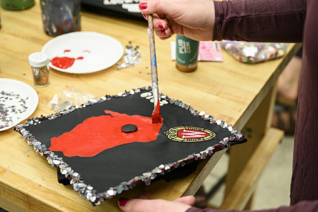 Photo: A hand holding a paintbrush paints a red state of Wisconsin on a cap.