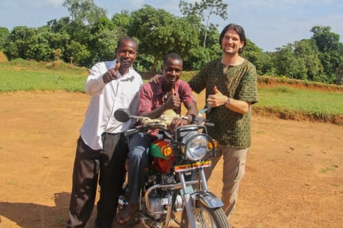 Photo: A man sits on a motorcycle, and two other people stand next to him, in Africa.