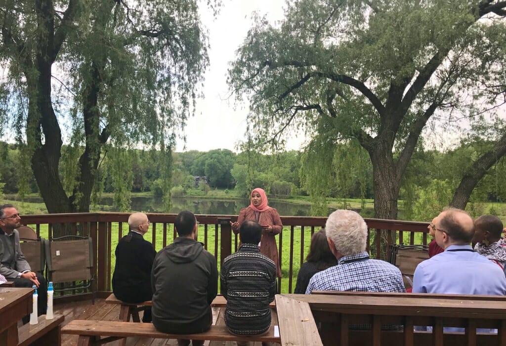Photo: Woman standing on deck speaking to seated people in front of pond and trees