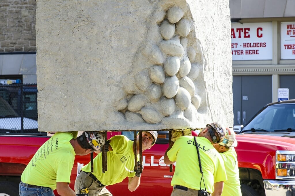 Photo: Four workers hold the statue under its base by their shoulders.