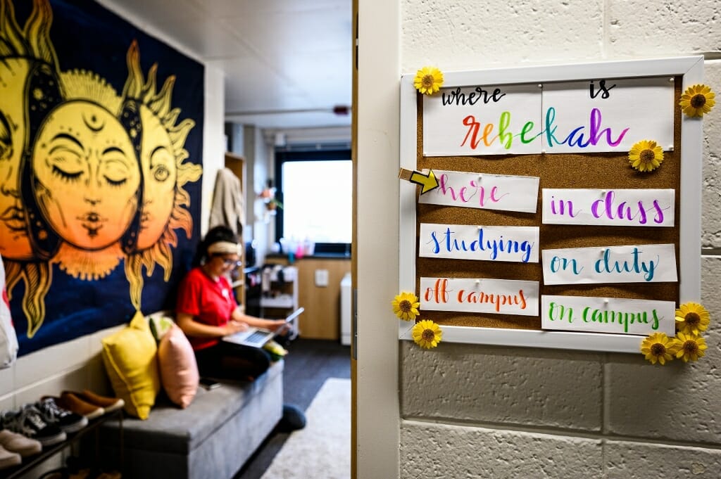 Photo of house fellow Rebekah Pulido seated on her bed; colorful signs on her door let residents know where she is.