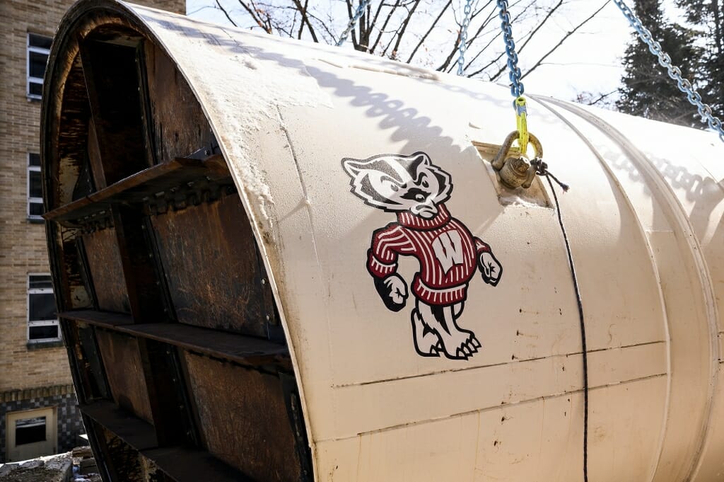 Construction workers prepare to lower by crane an 11-foot diameter tunnel bore head – adorned with a logo of Bucky Badger – down into a reinforced earthen pit to starts its work.