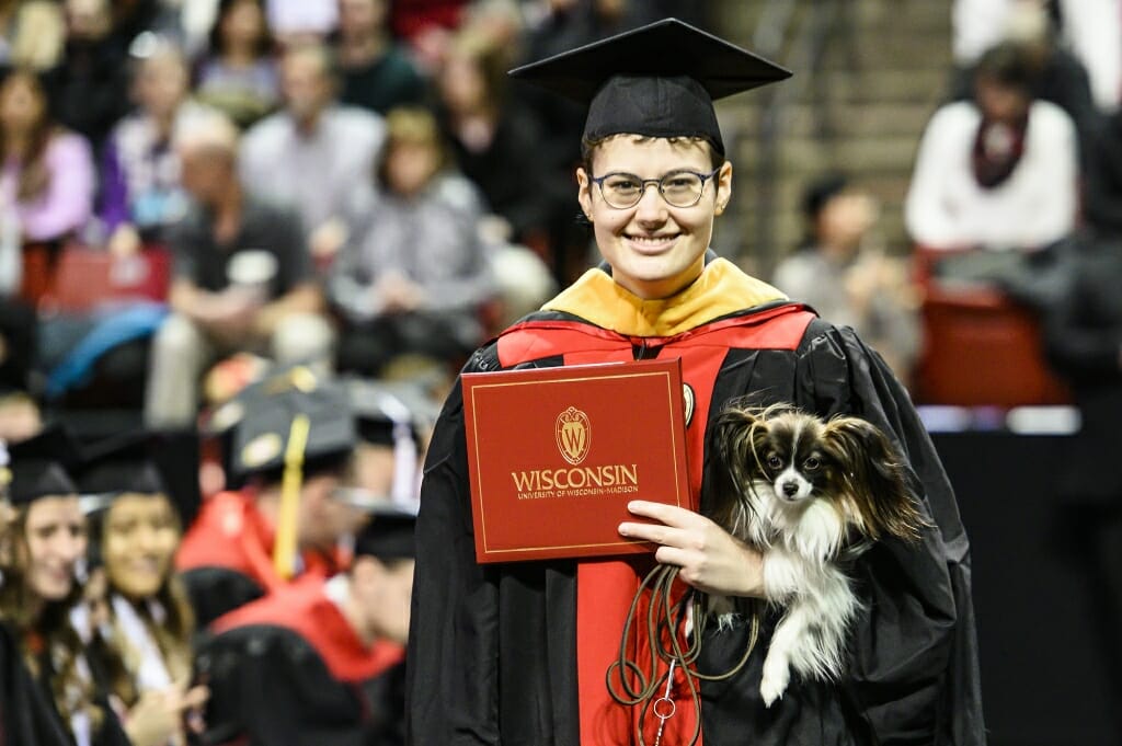 Photo of a graduate holding her small black-and-white service dog and her diploma cover.