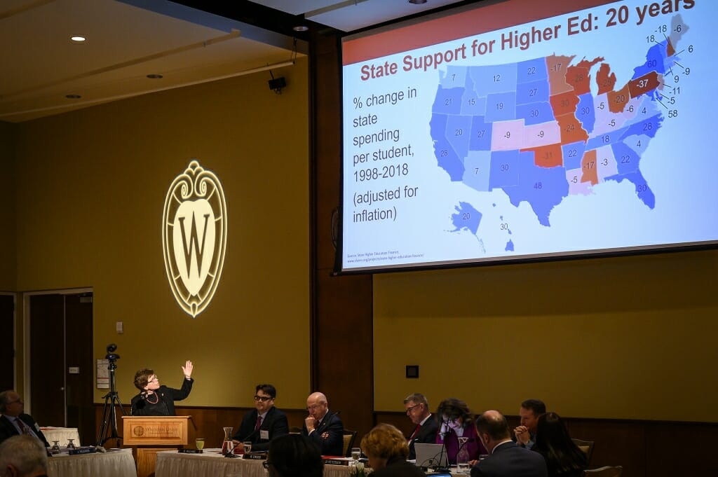 Rebecca Blank standing at a podium under a projected W crest, gesturing toward a map of U.S. states showing changes in higher ed support over 20 years. Wisconsin is in red, indicating a decrease.
