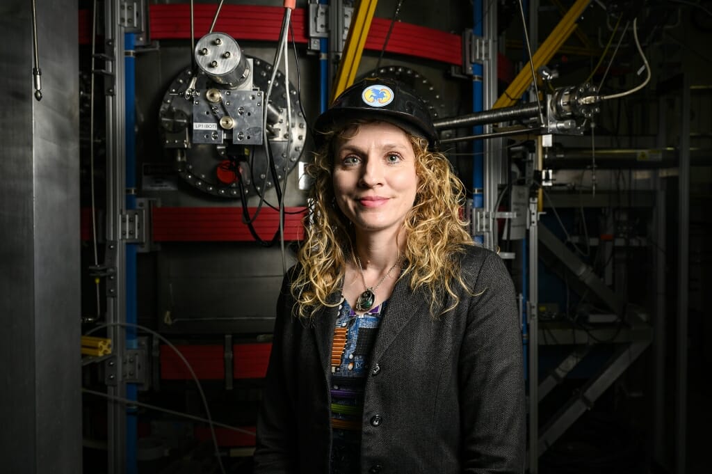 Steffi Diem standing in front of a round metal chamber with wires attached containing powerful magnets into which plasmas are injected. She is wearing a hat with a pegasus logo.