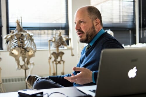 John Hawks sitting at his desk with a laptop open and hominid skeletons in background