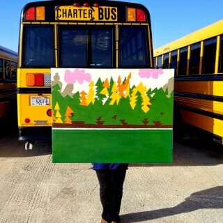 Pinc holding painting in front of yellow bus
