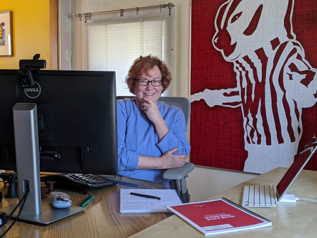 Linda Zwicker sitting opposite her computer on a desk with picture of Bucky Badger on wall behind her