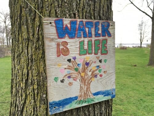 Sign the says WATER IS LIFE with a tree next to a waterway, hanging on a tree trunk