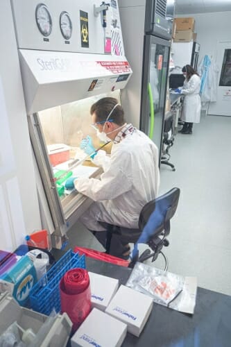A man in a white lab coat sits at a desk and uses lab equipment.