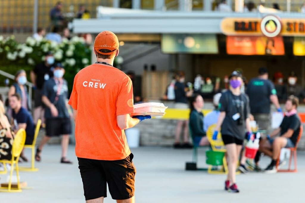 A man wearing a mask and an orange Wisconsin Union shirt carries food to a table.