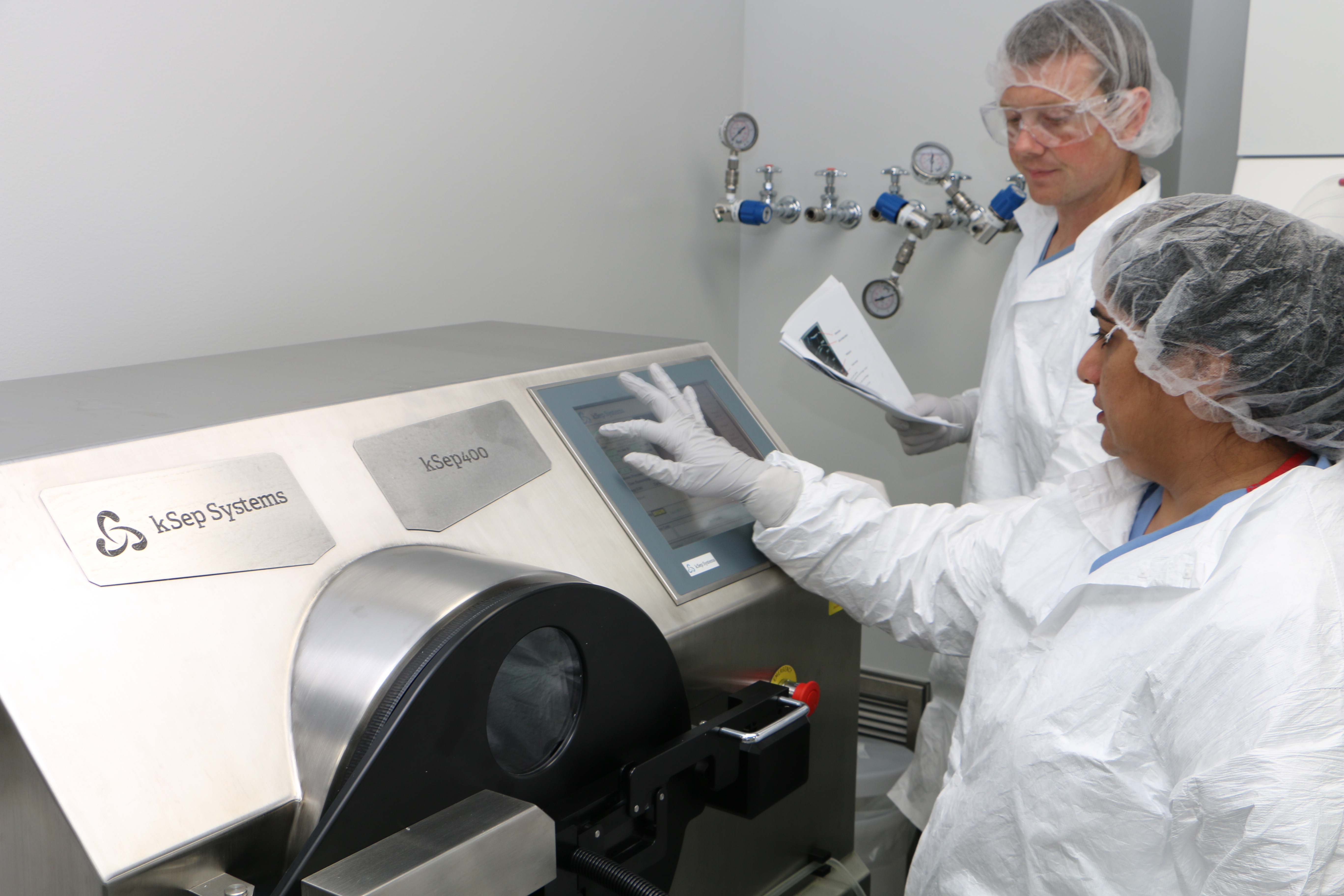 Photo of two staff members in protective lab gear standing next to a large centrifuge.