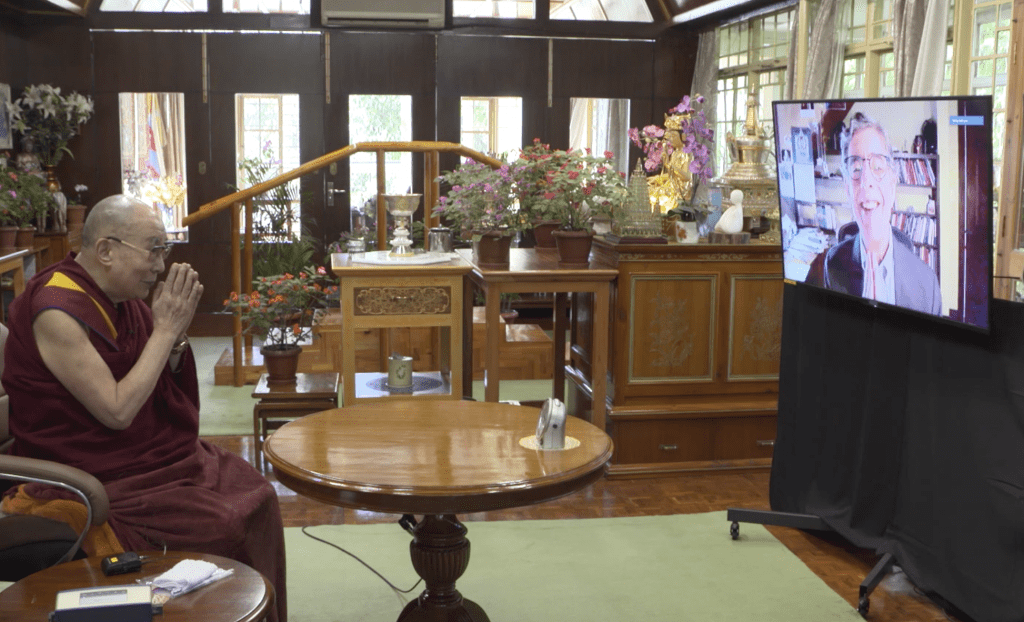 Dalai Lama sitting in a chair conversing with Richard Davidson on a video screen