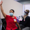 Photo of Tina Schubert waving her hand in celebration as she receives a vaccine injection in her arm.