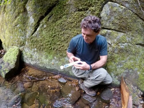 Tony crouching next to rocks and holding a test tube