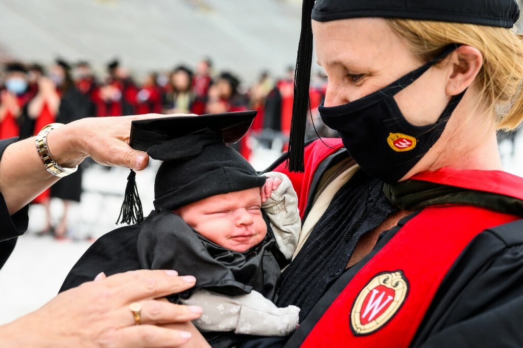 Photo of Hilton holding her baby to her chest as a friend adjusts the baby's mortarboard.