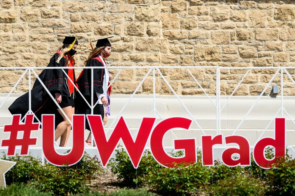 Photo of two women in caps and gowns walking past a giant sign spelling out "#UWGrad."