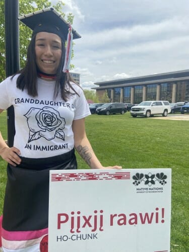 Photo of Marissa Fernandez wearing her mortarboard standing outside Gordon Dining & Events Center with a sign saying "congratulations" in Ho-Chunk.