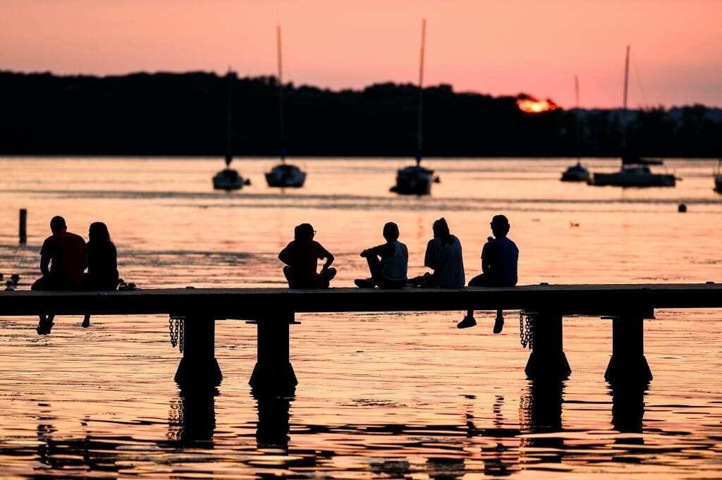 People are silhouetted by the setting sun as they sit along the lake.