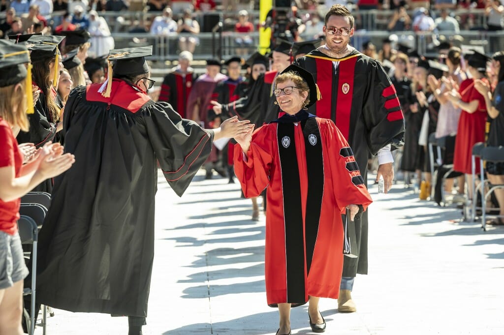 Chancellor Rebecca Blank, wearing red and black Wisconsin academic regalia, exchanges high fives with a graduate in cap and gown